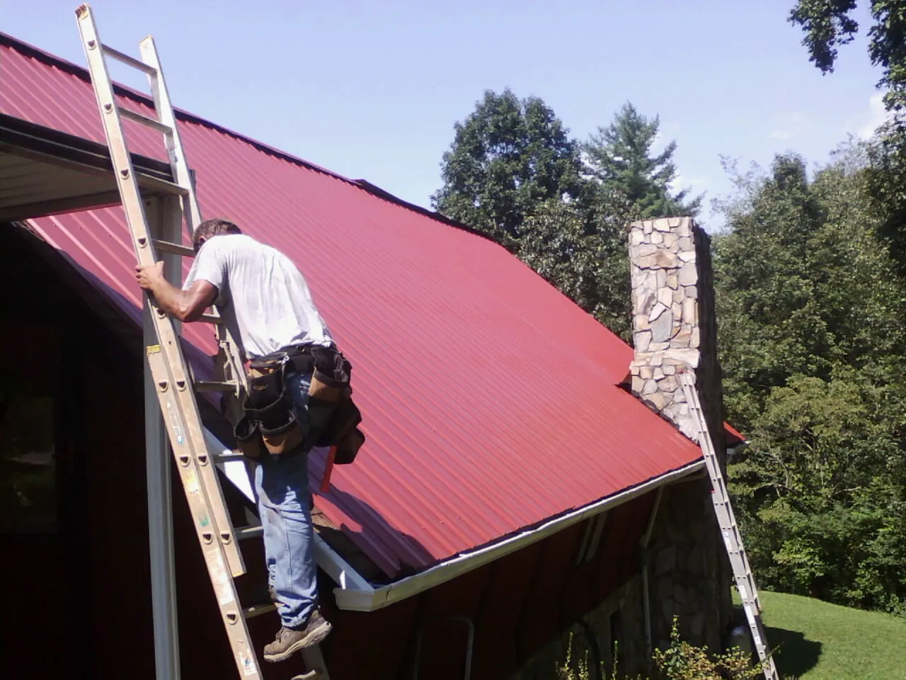 Roofer on a ladder beside a red metal roof and stone chimney — Soffit & Fascia Repair by Roofing Brothers in Rocky Mount